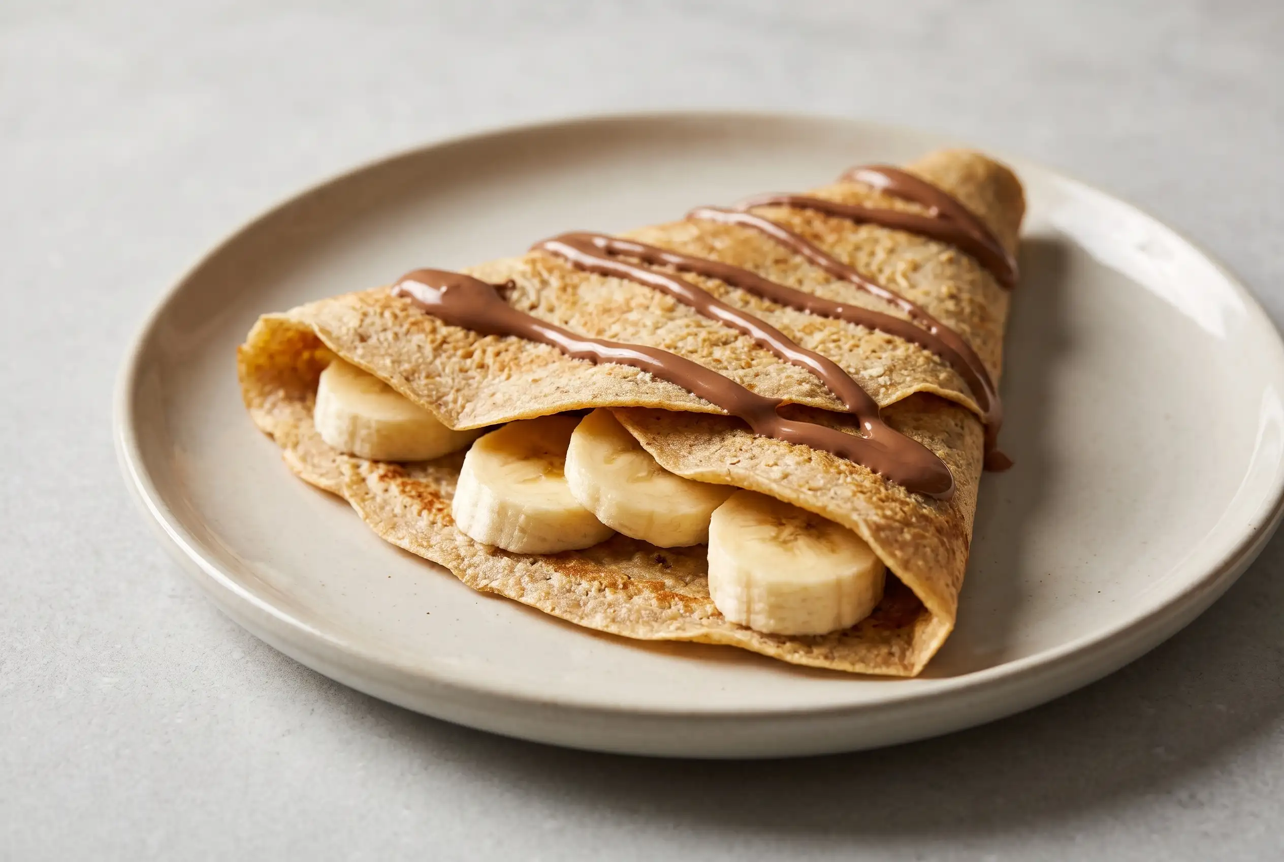 Almond oat crepe folded with banana slices and melted chocolate on a ceramic plate