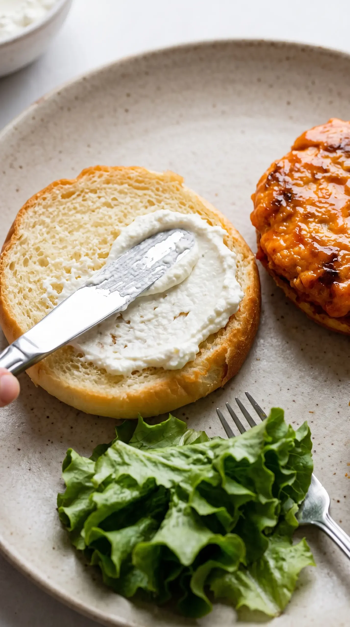 Whipped feta spread being added to a bun during buffalo chicken burger assembly